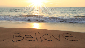 The word “Believe” written in the sand at a beach with waves in the ocean as the sun shines on a beautiful day.