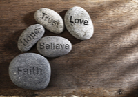 Inspirational text with the words “Faith, Trust, Love, Hope, and Believe” written on pebbles on a wooded backdrop.