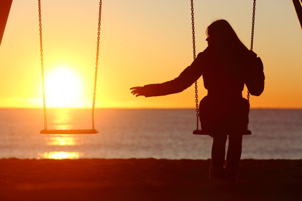 A woman sits alone next to an empty swing at the beach as the sun sets. Concept - God sits with you.