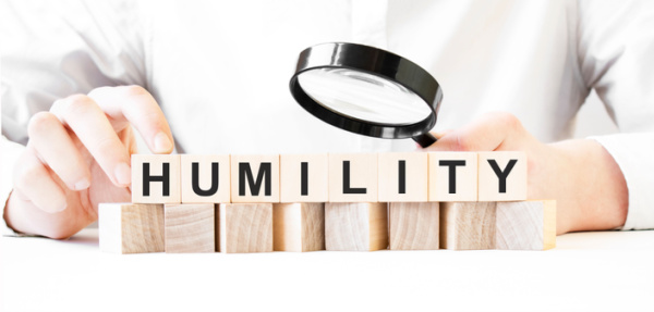 A businessperson uses a magnifying glass to examine a wooden block with the word "HUMILITY" carved into it.