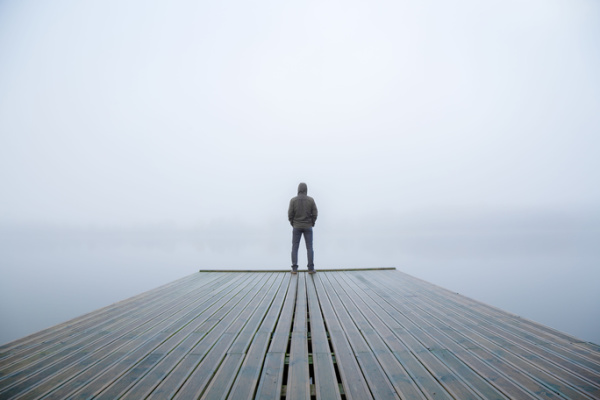 A young man stands alone on the tip of a dock, staring at a lake early on a chilly autumn morning. A foggy atmosphere surrounds him, and he’s looking over the water. The scene is filled with beauty and freedom, depicting a moment of peace outdoors. Concept - When God Isolates You.