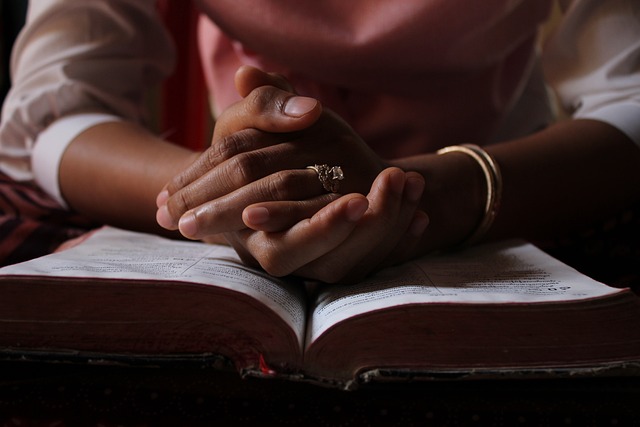 Hands are pictured with fingers interlaced, forming a gesture of prayer on a Bible.