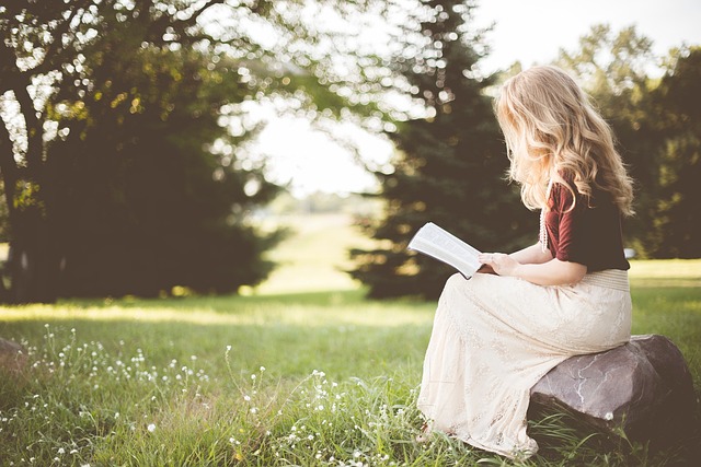 A woman sits on a boulder with grass beneath it and three trees in the background, reading her bible. Concept, meditating on God's Word.