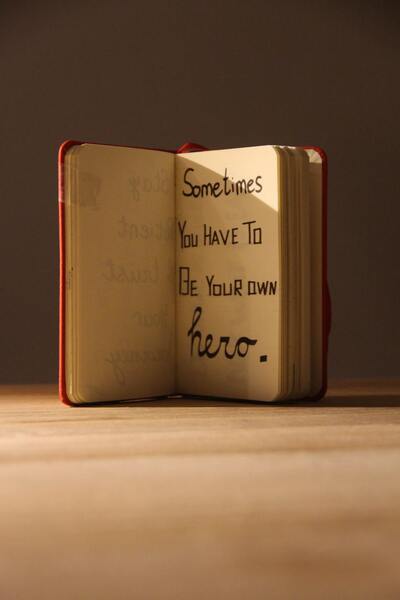 A book sitting on a wooden desk with the phrase "Be your own hero" written across the pages.