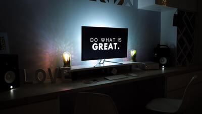 On a desk with the word "LOVE" carved out of wood sits a computer with the phrase "Do What Is Great" on the screen. Two candles sit on either side of the computer, dimly illuminating the room.