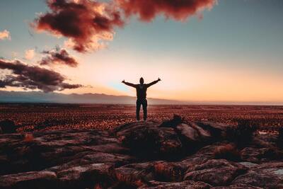 A man standing on a boulder in an open desert with billowing clouds and a sunrise is pictured with his arms raised.