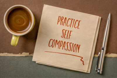 A napkin containing the words' practice self-compassion handwritten with a coffee cup and pen.