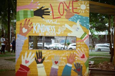 In the backdrop of a scene featuring trees and cars, there is a signpost that displays a collection of love and kindness words in the form of a collage. The collage signpost is surrounded by hands pointing toward it, emphasizing its message.