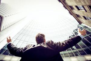 A man who appears to be a businessman stands amidst a group of buildings and stretches his arms toward the sky.