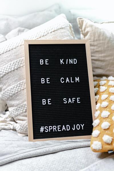 On a couch, there are throw pillows surrounding a chalkboard that displays the message "Be Kind, Be Calm, Be Safe, Spread Joy." Concept, spreading love and kindness.