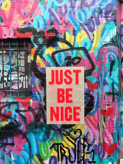 A brick wall displays an abstract painting that includes the colors red, blue, and yellow. The artwork features the phrase "Just Be Nice" in the center of the painting. Concept, spreading love and kindness.